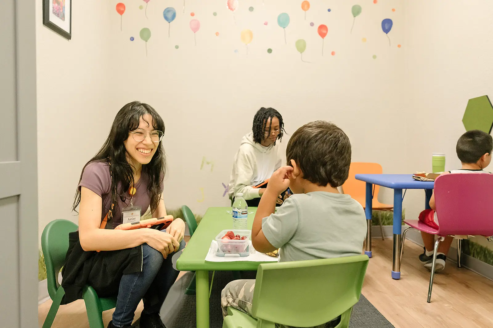 Cheerful therapist interacts with child in colorful room. Balloon wall decals, diverse children, and nutritious snacks create welcoming atmosphere