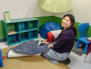 Smiling ABA Professional in colorful child-friendly room with child playing. Blue furniture, storage cubbies, and soft elements create inviting space