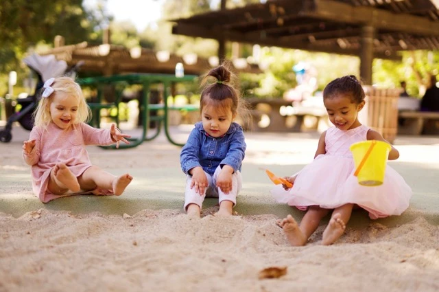 Three happy toddlers playing in a sandbox at a park, smiling and using toys